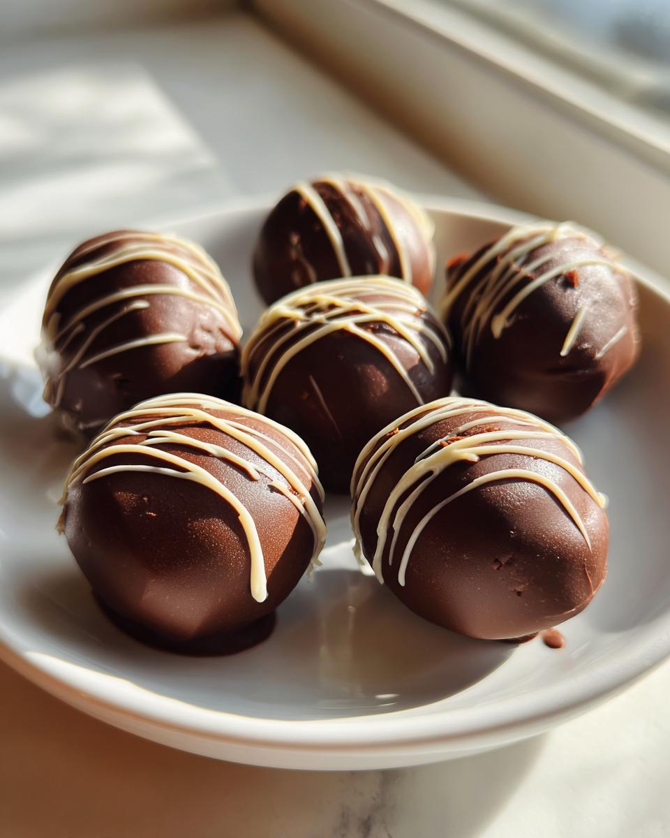 Six chocolate covered oreo balls drizzled with white chocolate, resting on a white plate in sunlight.
