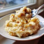 A fork lifting a generous bite of fluffy garlic mashed potatoes topped with browned bits from a white bowl.