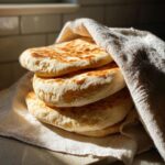 A stack of four golden-brown, freshly cooked pita bread pockets wrapped partially in a white linen cloth.