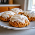 Four soft, orange pumpkin cookie treats topped with thick white cream cheese frosting on a light gray plate.