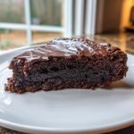 A rich, fudgy slice of flourless chocolate cake topped with melted chocolate ganache, sitting on a white plate.