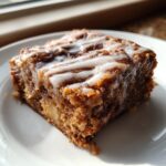 A single square of moist, dark apple brownies topped with a thick white glaze, served on a white plate.