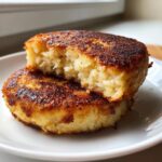 Two golden brown, pan-fried mashed potato cakes stacked, with the top one broken open to show the fluffy interior.