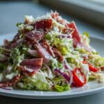 A close-up, tempting view of a fully assembled grinder salad featuring chopped lettuce, salami, red onion, tomatoes, and creamy dressing.
