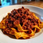 Close-up of hearty bolognese sauce generously spooned over wide, fresh egg pasta ribbons in a white bowl.
