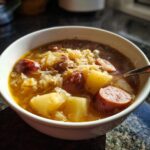 A steaming white bowl filled with rich sauerkraut soup featuring chunks of potato and sliced smoked sausage.
