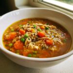 Close-up of a white bowl filled with rich turkey soup featuring shredded turkey, wild rice, bright orange carrots, and green peas.