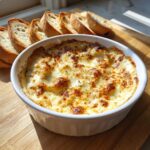 A bubbling, hot cheesy bread dip served in a white ramekin next to slices of toasted baguette.