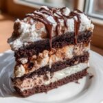 A tall slice of rich, layered hot chocolate poke cake featuring dark chocolate cake, white and brown fillings, topped with whipped cream and chocolate drizzle.