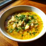 Close-up of a white bowl filled with vibrant yellow Lemon Ginger Turmeric Chicken and Rice Soup, topped with fresh parsley.