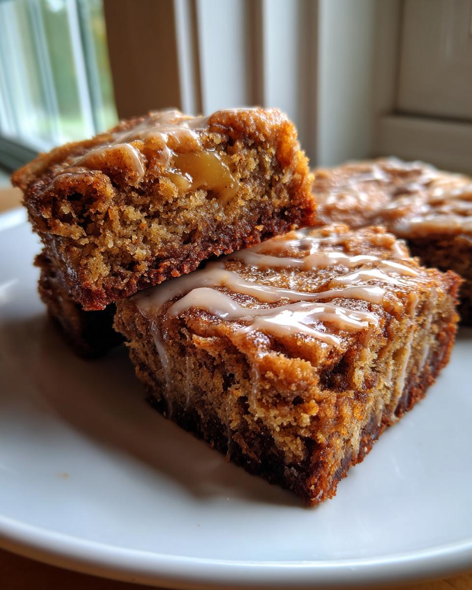 Close-up of moist apple brownies stacked on a white plate, drizzled with a light cinnamon glaze.