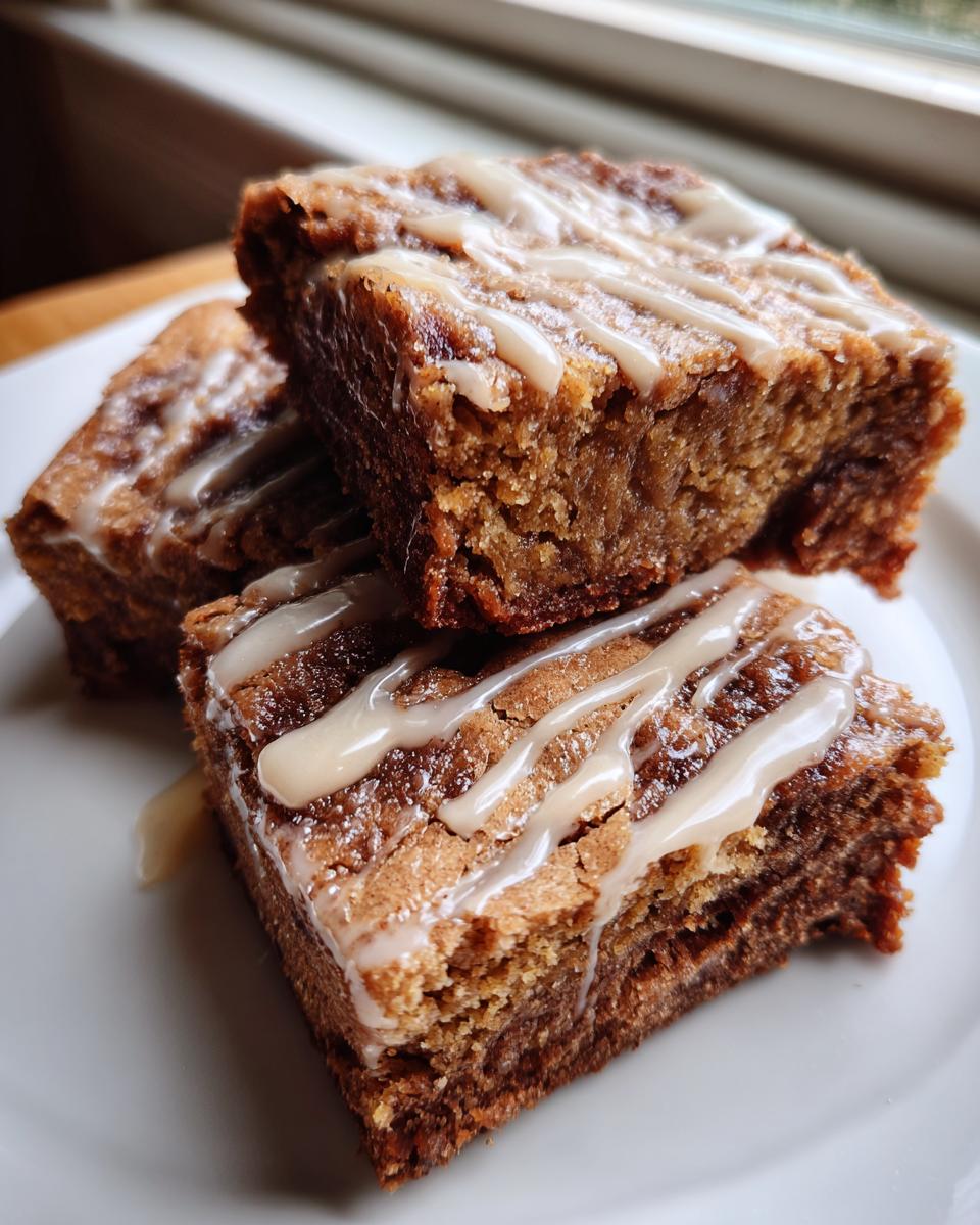 Three moist apple brownies stacked on a white plate, drizzled with a thick vanilla glaze.