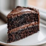 Close-up of a rich, dark slice of moist chocolate cake layered with thick chocolate frosting.