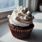 A close-up of a moist hot chocolate cupcake topped with white whipped frosting and mini marshmallows.