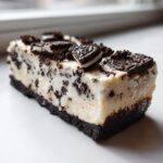 A close-up side view of a rich slice of Oreo cheesecake with a dark cookie crust and cream filling loaded with cookie pieces.