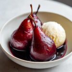 Close-up of three glossy, deep red poached pears served in a white bowl with a scoop of vanilla ice cream.