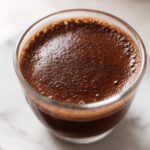 Close-up of rich, dark homemade chocolate pudding with a foamy top in a clear glass.