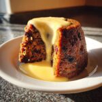 A close-up of a rich, dark plum pudding slice generously topped with flowing vanilla custard.