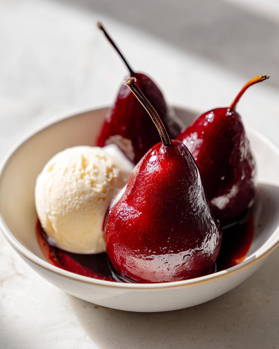 Three glossy, deep red poached pears served in a white bowl next to a scoop of vanilla ice cream.