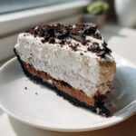 A decadent slice of multi-layered oreo pie with a dark crust, chocolate filling, and whipped topping.