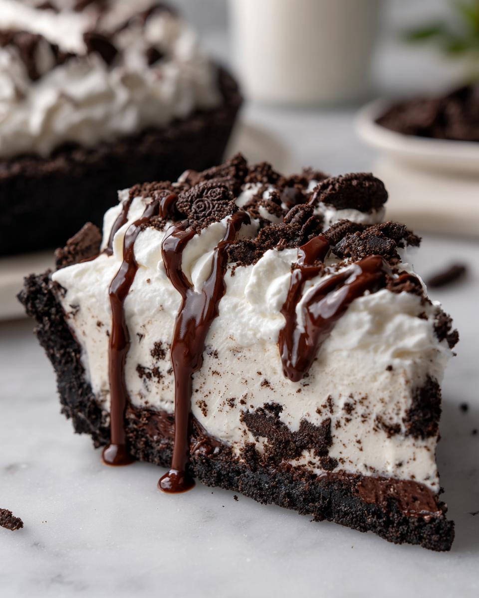 A close-up slice of rich ice cream pie featuring a dark cookie crust, creamy filling, whipped topping, and chocolate syrup drizzle.