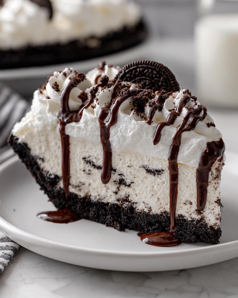 A decadent slice of ice cream pie featuring a dark cookie crust, creamy filling, whipped topping, and chocolate syrup drizzle.