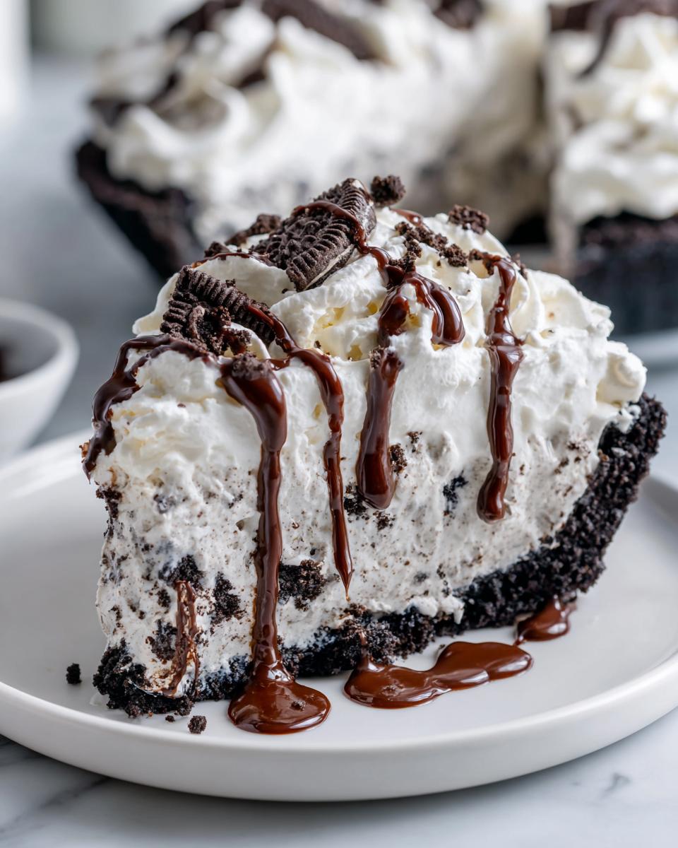 A decadent slice of ice cream pie featuring a dark cookie crust, creamy filling with cookie chunks, topped with whipped cream and chocolate sauce.