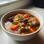 Close-up of a white bowl filled with rich beef barley soup, featuring chunks of beef, carrots, celery, and barley.