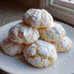 A stack of soft, powdered sugar-dusted cool whip cookies resting on a white plate near a window.