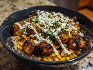 A close-up of a street corn chicken rice bowl topped with charred chicken, roasted corn, cotija cheese, and crema.