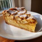 A moist slice of swedish apple cake topped with baked apple slices and dusted with powdered sugar on a white plate.