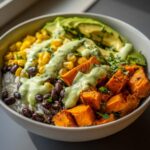 A vibrant sweet potato taco bowl featuring roasted sweet potatoes, black beans, corn, rice, avocado slices, and a creamy green dressing.
