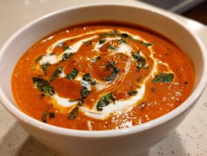 Close-up of a bowl of rich orange tomato basil soup, swirled with cream and topped with fresh basil.
