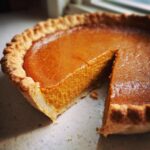 A close-up of a freshly baked vegan pumpkin pie with one slice partially removed, showing the creamy orange filling.
