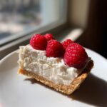 A close-up of a slice of white chocolate mousse tart with a graham cracker crust, topped with creamy filling and fresh raspberries.