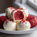 A stack of decadent red velvet cake balls coated in white chocolate, one cut open to show the vibrant red interior.