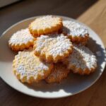 A stack of golden 3-ingredient butter cookies dusted heavily with powdered sugar on a white plate.