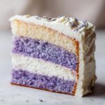 Close-up of a tall slice of amazing lavender cake showing alternating purple and white layers, topped with white frosting and dried lavender buds.
