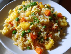 A close-up view of vibrant pineapple fried rice featuring chunks of pineapple, carrots, peas, egg, and green onions on a white plate.