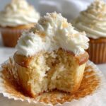 Close-up of a banana pudding cupcakes with a bite taken out, showing moist cake and creamy frosting topped with crumbs.