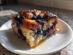 A close-up, sunlit serving of Blueberry French Toast Casserole on a white plate, showing caramelized edges and moist interior.