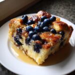 A close-up of a square slice of moist Blueberry French Toast Casserole topped with fresh blueberries and drizzled with syrup.
