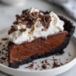 A close-up of a decadent slice of chocolate mousse pie with a dark crust, topped with whipped cream and chocolate shavings.