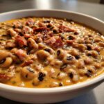 Close-up of creamy black eyed peas in a white bowl, topped with crispy bacon pieces.