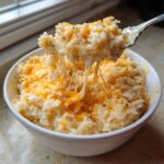 A spoonful of creamy, cheesy rice showing long cheese pulls from the bowl of cheesy rice.