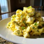 A close-up of a mound of creamy, yellow egg salad recipe topped with chopped green onions on a white plate.