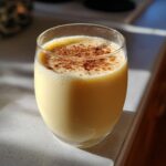 A close-up of a glass filled with creamy, pale yellow eggnog, topped with a dusting of cinnamon.