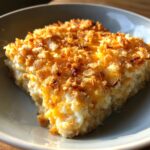 A close-up of a square serving of creamy rice casserole topped with melted cheese and crunchy, golden-brown cornflakes.