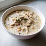 A close-up of creamy white chicken soup topped with shredded white chicken, white beans, and fresh thyme.