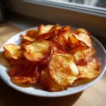A close-up of freshly made, golden brown potato chips seasoned with paprika, piled high on a white plate.